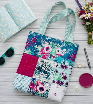 Colorful patchwork tote bag on a wooden surface with a notebook, sunglasses, and flowers.