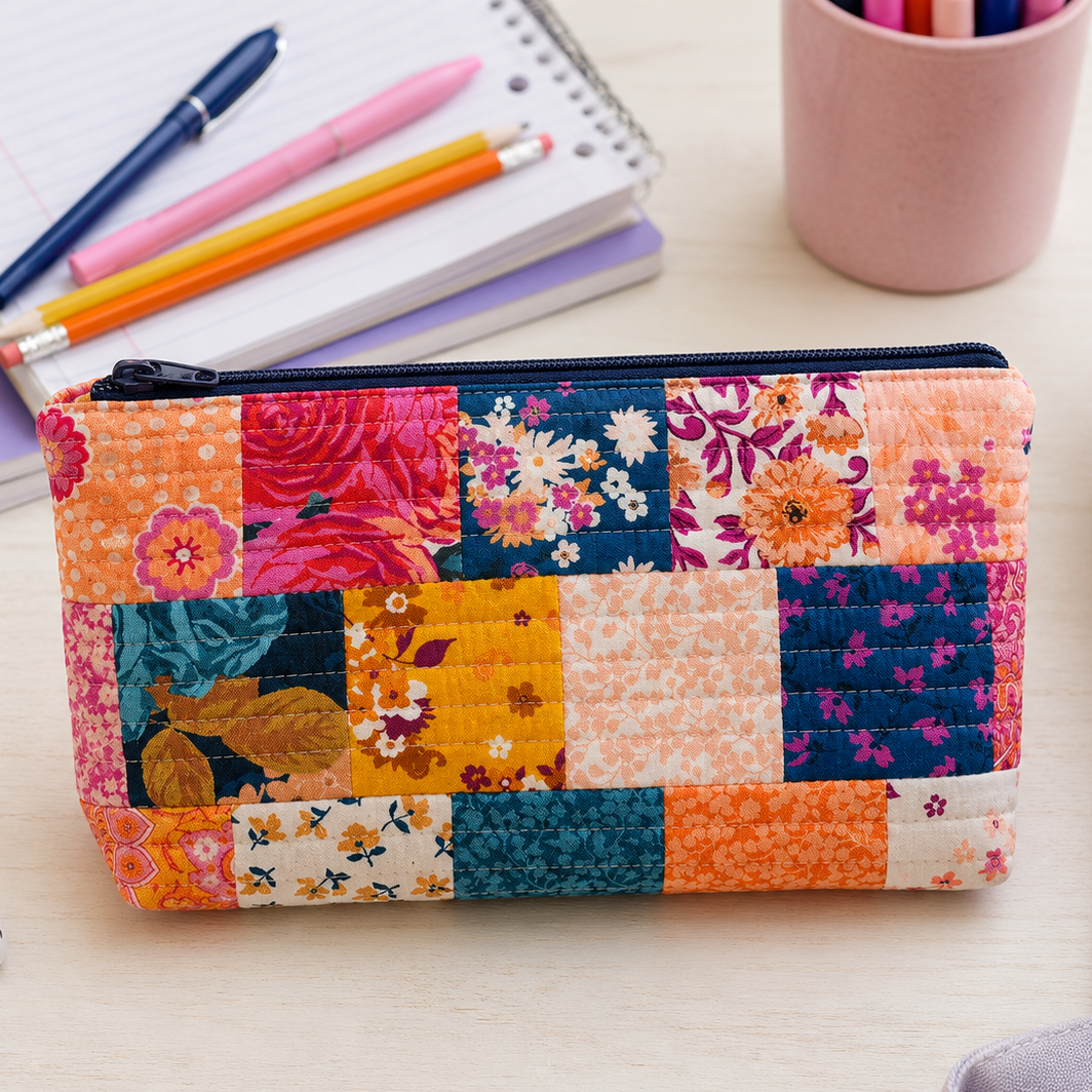 A rectangular patchwork quilted pouch with a floral and geometric print in orange, pink, blue, yellow, and cream, shown on a light wood desk with notebooks, pens, earbuds, and a lavender bag nearby.