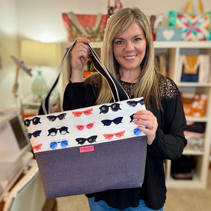 Woman holding a colorful tote bag with cat designs in a room with shelves in the background.