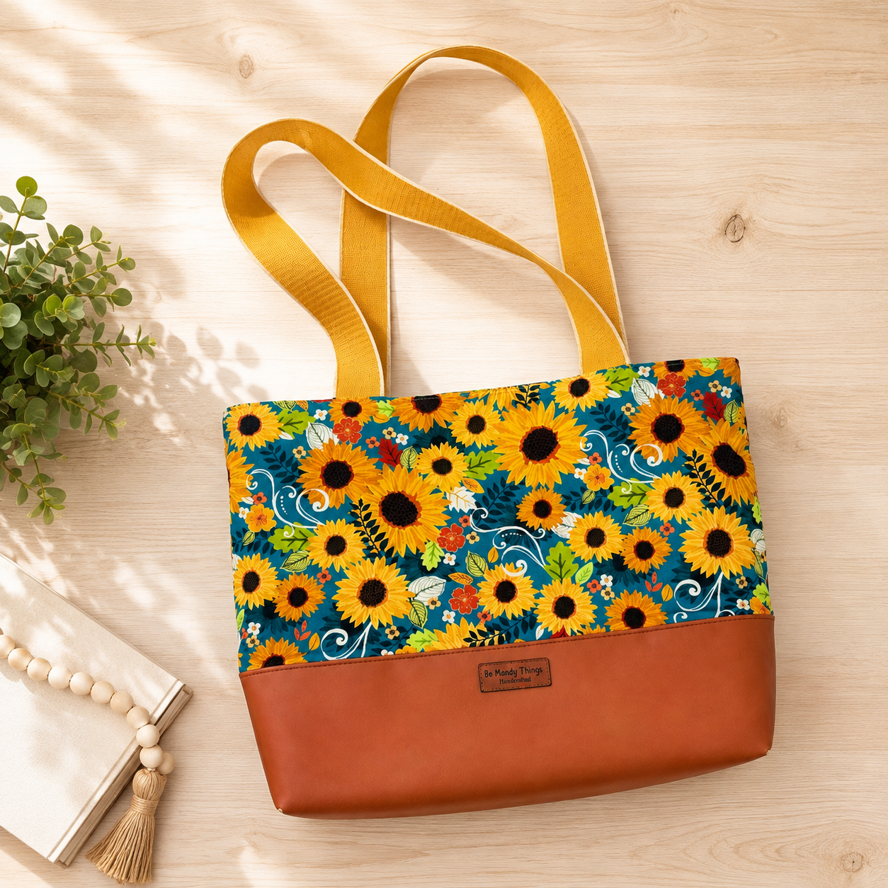 Sunflower-print Joanna tote bag with mustard-yellow handles and a warm brown faux-leather base, styled flat on a light wood surface with greenery and a neutral book.