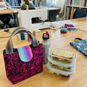 Table with a colorful bag, lunch container, and water bottle in a room with sewing equipment and bookshelves.