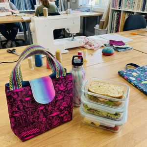 Table with a colorful bag, lunch container, and water bottle in a room with sewing equipment and bookshelves.