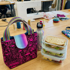 Table with a colorful bag, lunch container, and water bottle in a room with sewing equipment and bookshelves.
