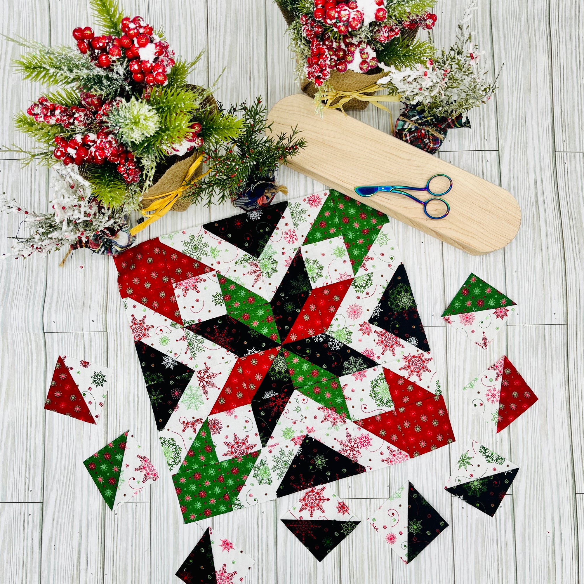 Star quilt block pattern in red green black and white fabric pieces arranged on a wooden floor with floral decorations and scissors