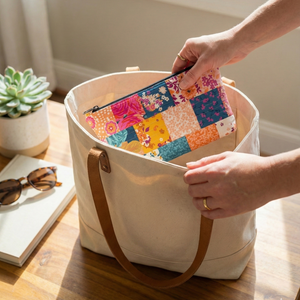 Person holding a colorful patchwork pouch over a large beige tote bag on a wooden surface.