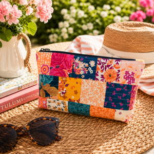 Colorful patchwork Frankie pouch on a woven surface with books, sunglasses, and flowers in the background.